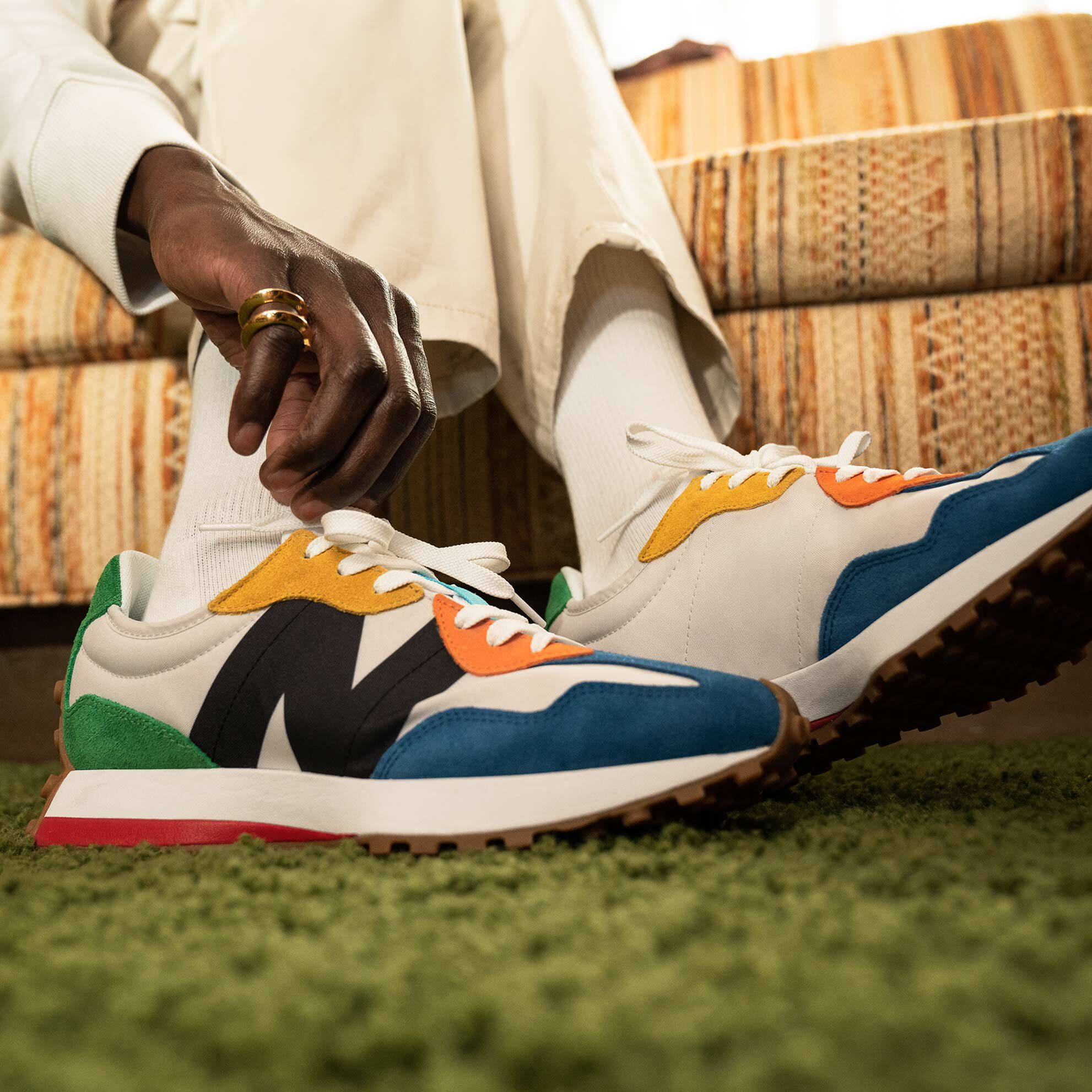 Close up crop of a pair of multicolor NB 327 sneakers worn by a man sitting on a couch in a 1970s décor room