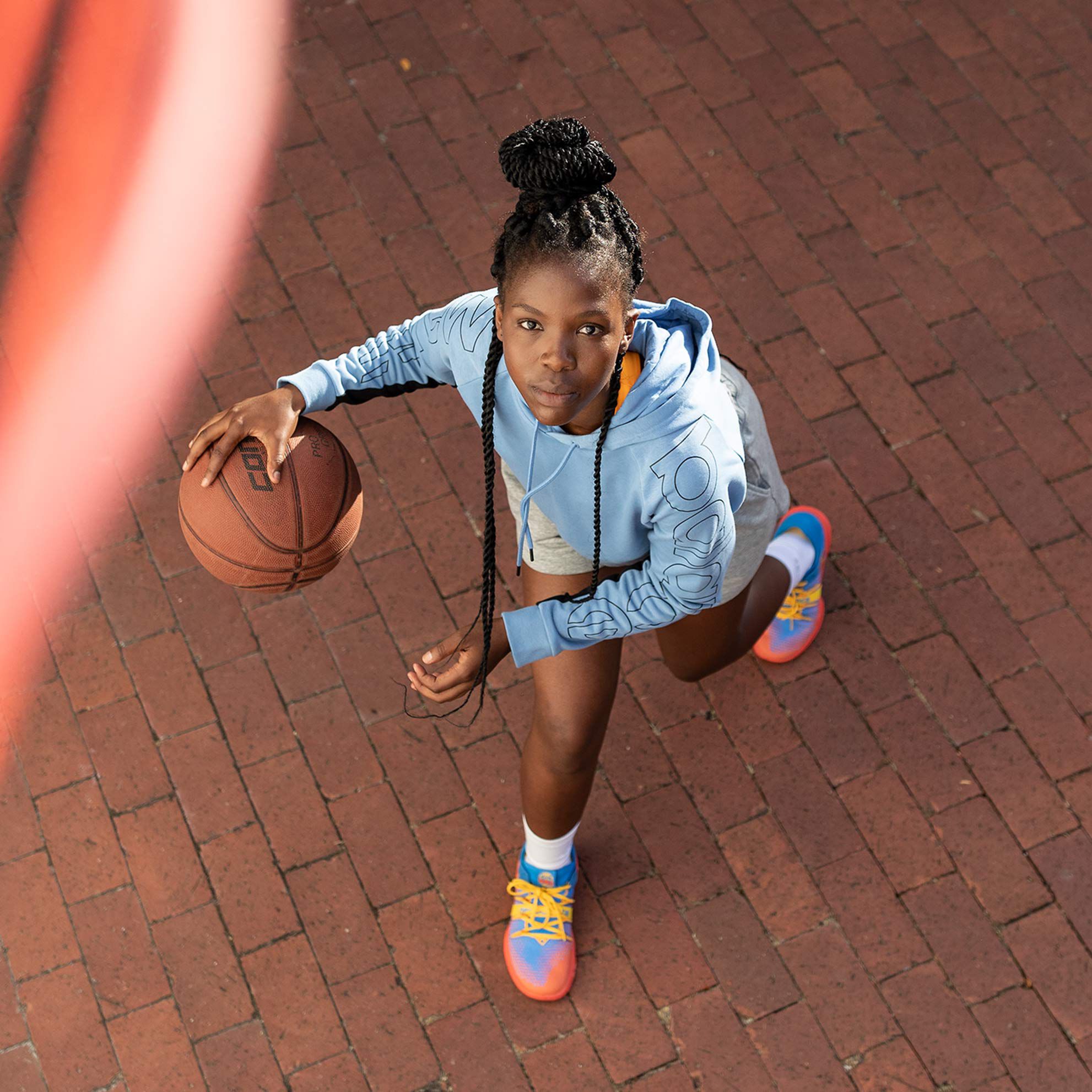 A teenage girl wearing a light blue NB hoodie and neon colored NB OMN1S Low basketball shoes as she approaches the hoop for a layup in her driveway