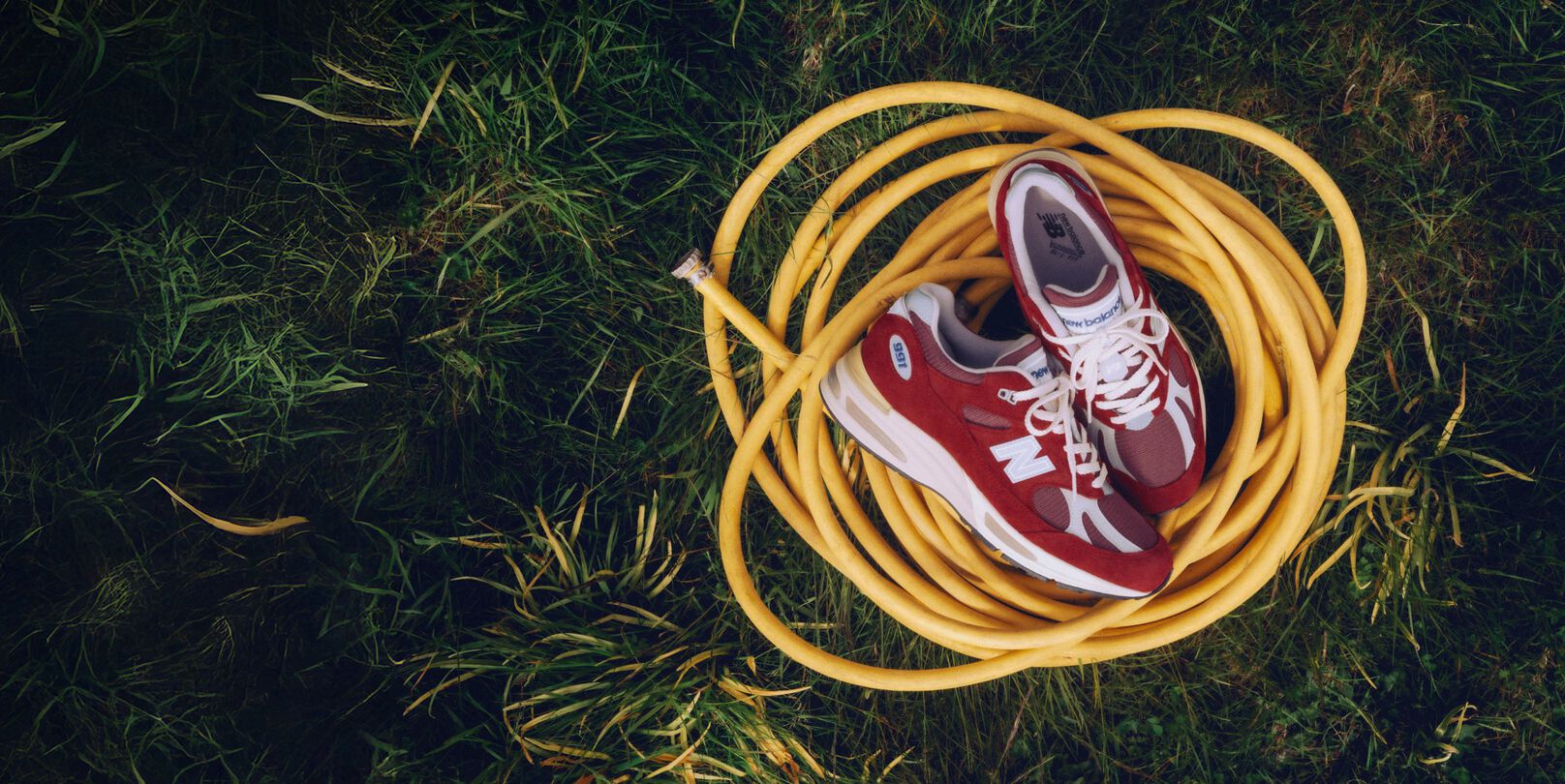 Une paire d'espadrilles New Balance Made in UK pos&eacute;es sur un tuyau jaune enroul&eacute;, au milieu d'une pelouse verdoyante.&nbsp;