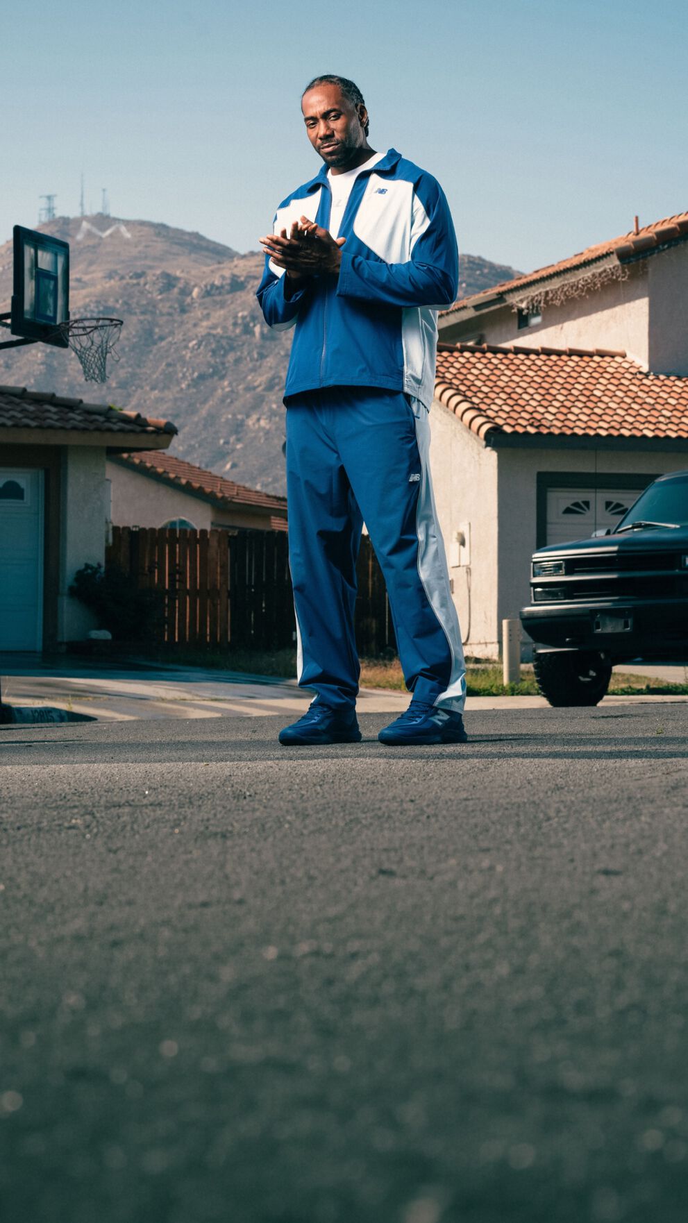 Une personne v&ecirc;tue d&rsquo;un ensemble de surv&ecirc;tement bleu et blanc et de chaussures de sport assorties se tient dans une rue de banlieue, avec des maisons et des montagnes en arri&egrave;re-plan.