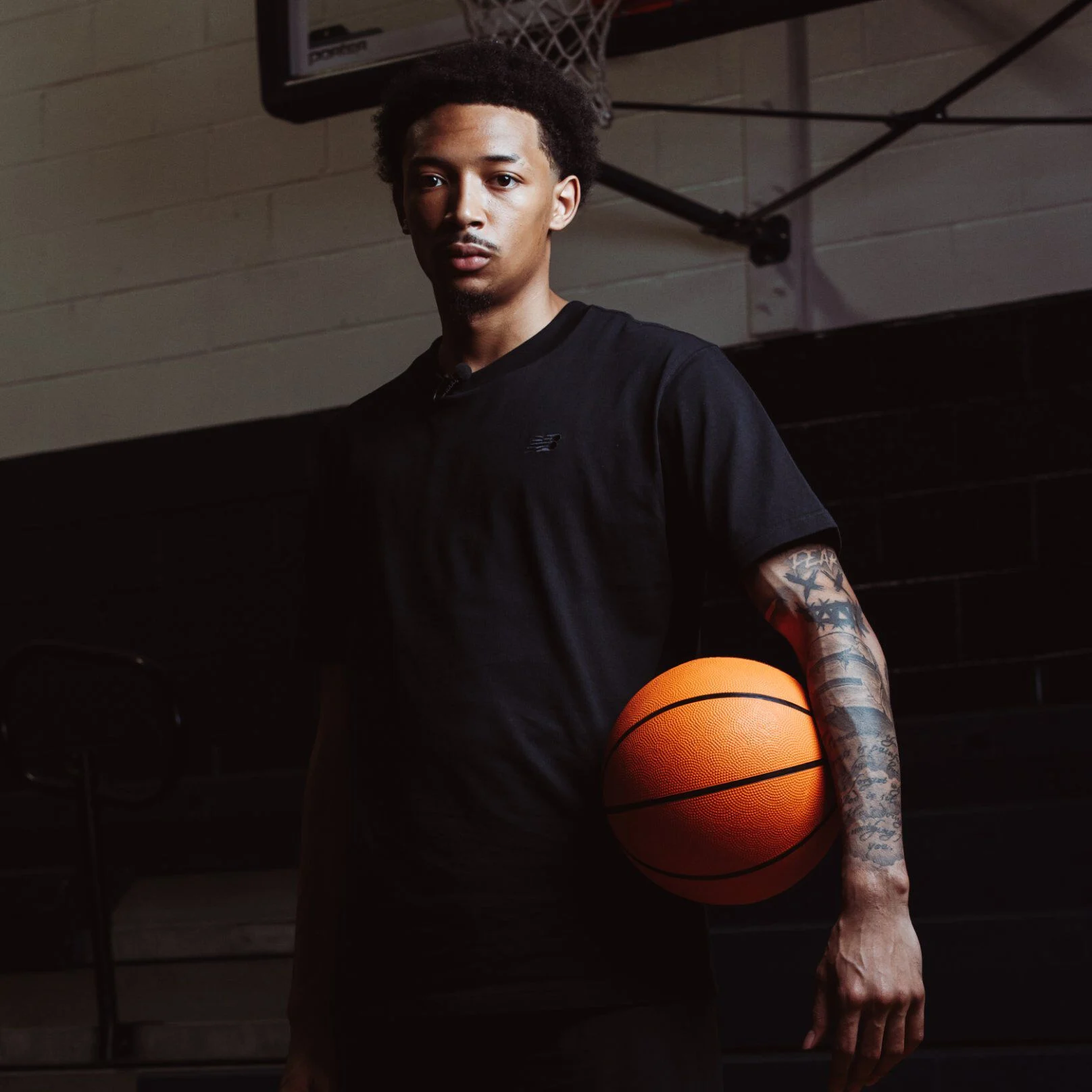 Une personne se tient sur un terrain de basket int&eacute;rieur, portant un t-shirt noir et tenant un ballon de basket contre sa hanche.