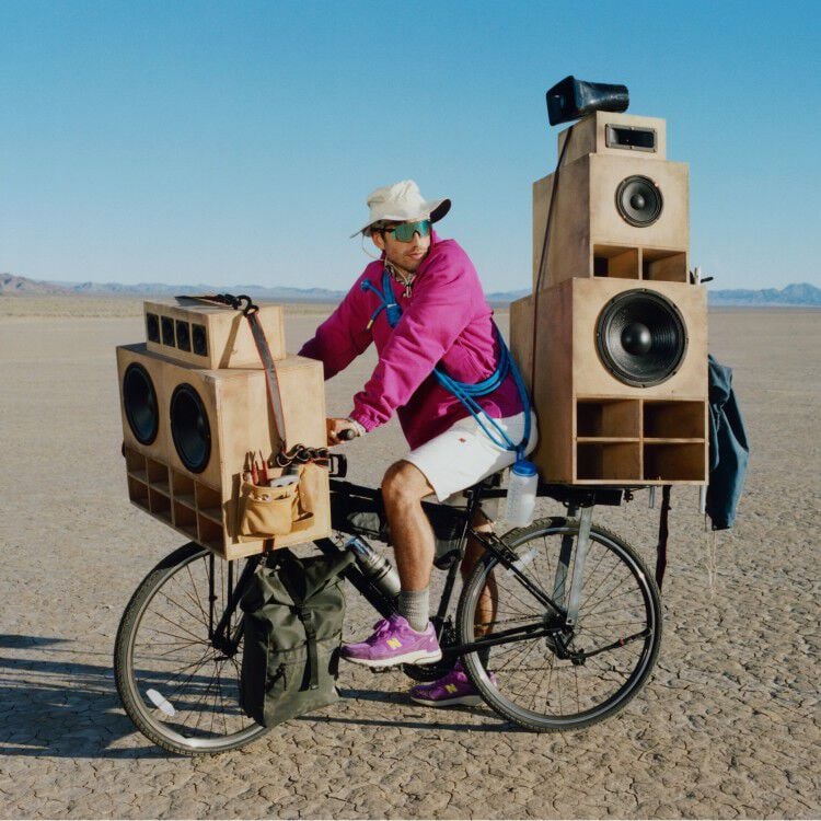 Guy on a bike with giant speakers attached to it wearing a purple sweater and shoes.