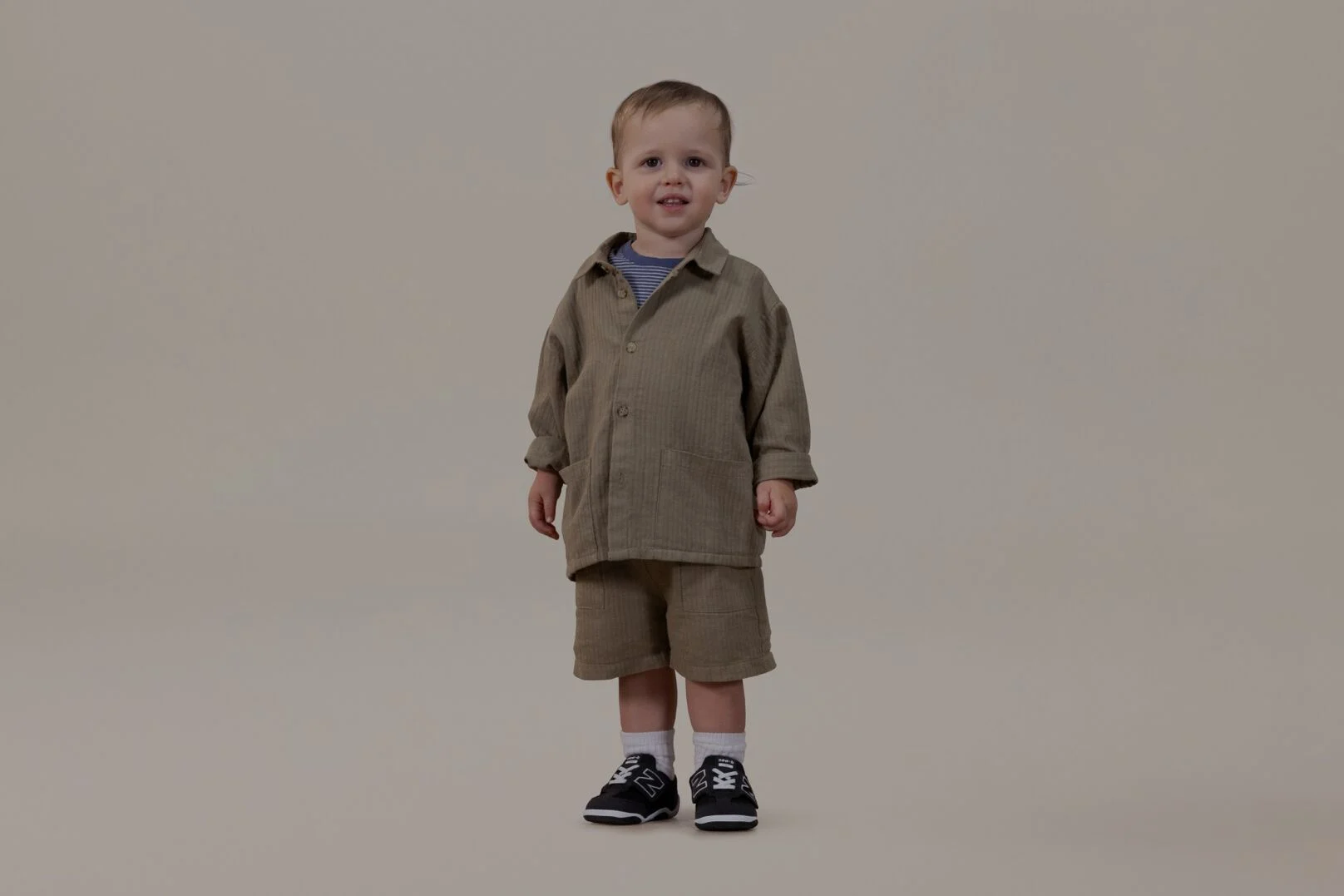 Little baby in a tan outfit standing on a grey background.