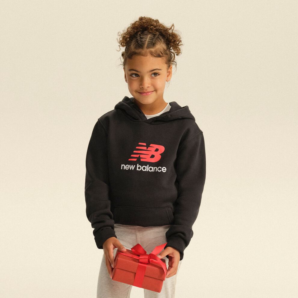 Enfant portant un chandail &agrave; capuchon New Balance noir et un pantalon d&rsquo;entra&icirc;nement gris, et tenant une bo&icirc;te-cadeau rouge dans un d&eacute;cor de studio &eacute;pur&eacute;.