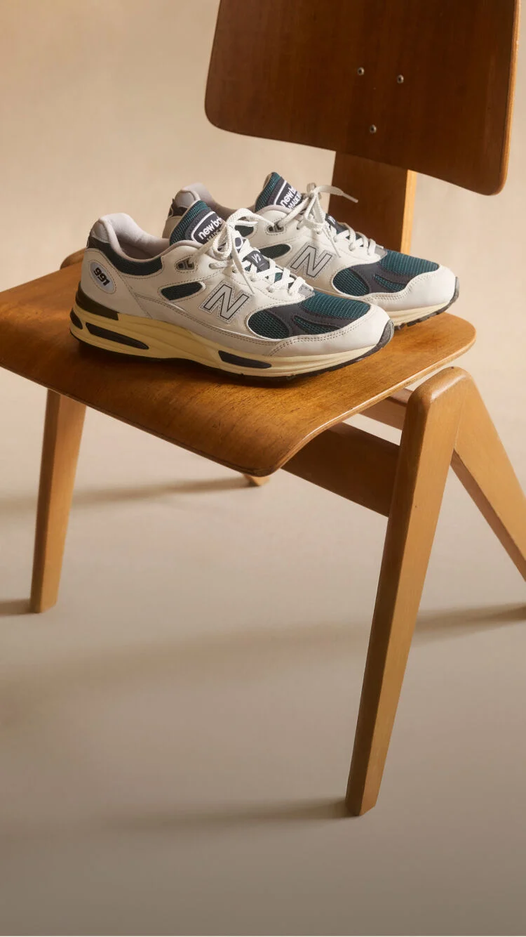 A pair of New Balance Made in UK sneakers sitting on a wooden chair in a beige studio environment. 