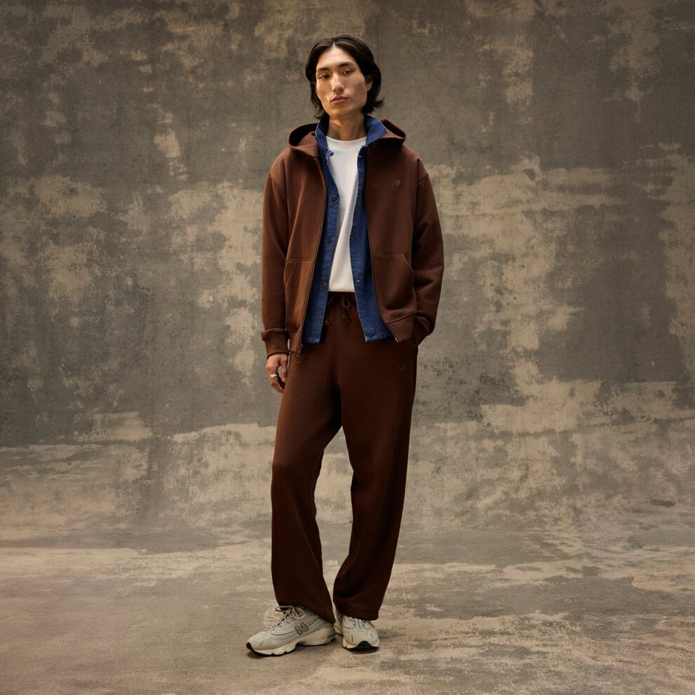 Person standing indoors against a textured backdrop, wearing a brown tracksuit layered over a blue inner piece and sneakers. 