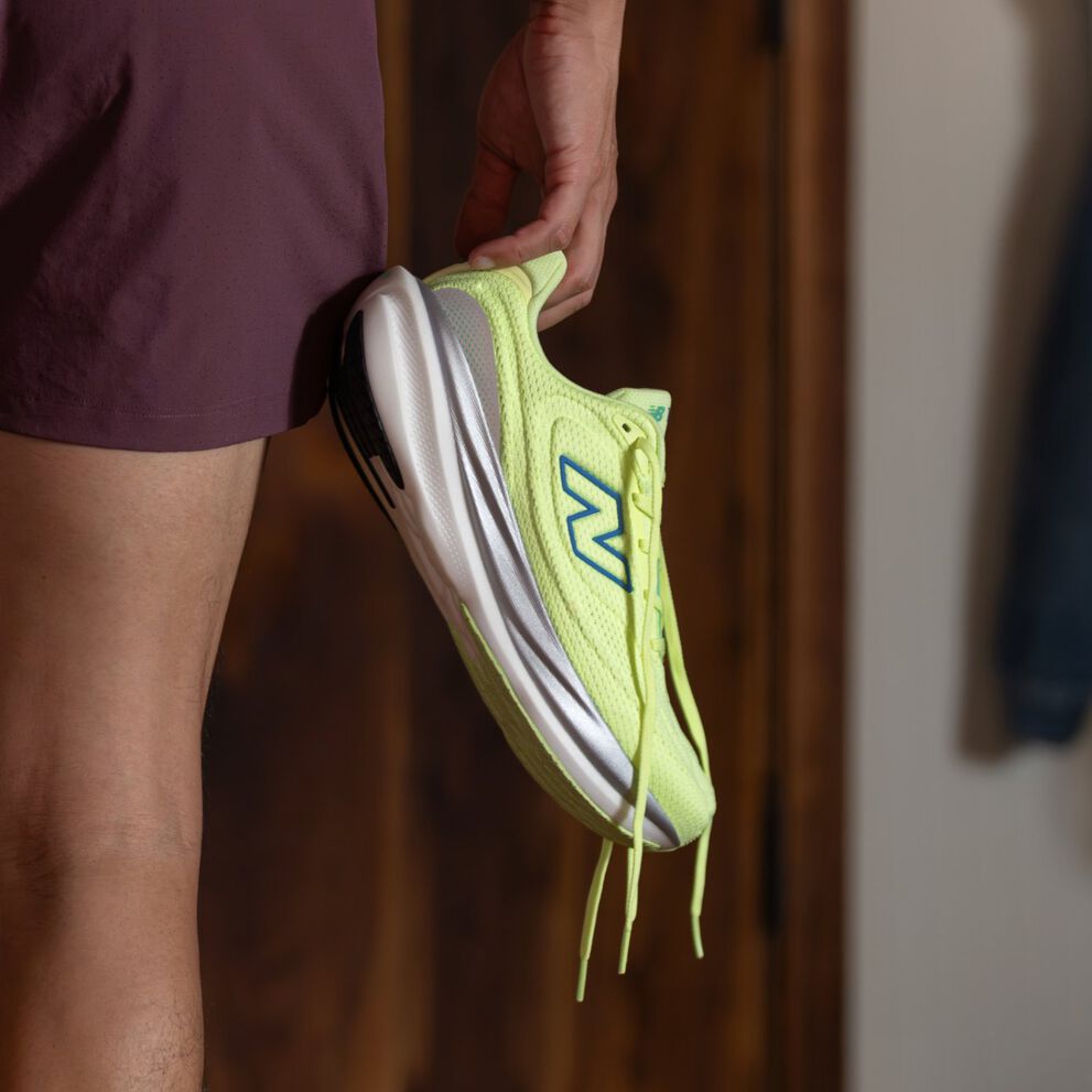 Gros plan d&rsquo;une chaussure de course New Balance jaune-vert vif avec logo bleu et semelle blanche, tenue par une personne portant un short bordeaux.&nbsp;