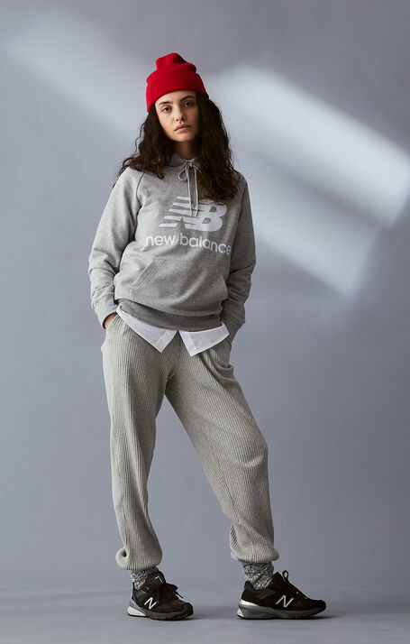 Woman posing wearing a grey NB Essentials hoodie with a red beanie and black NB 990v5 sneakers