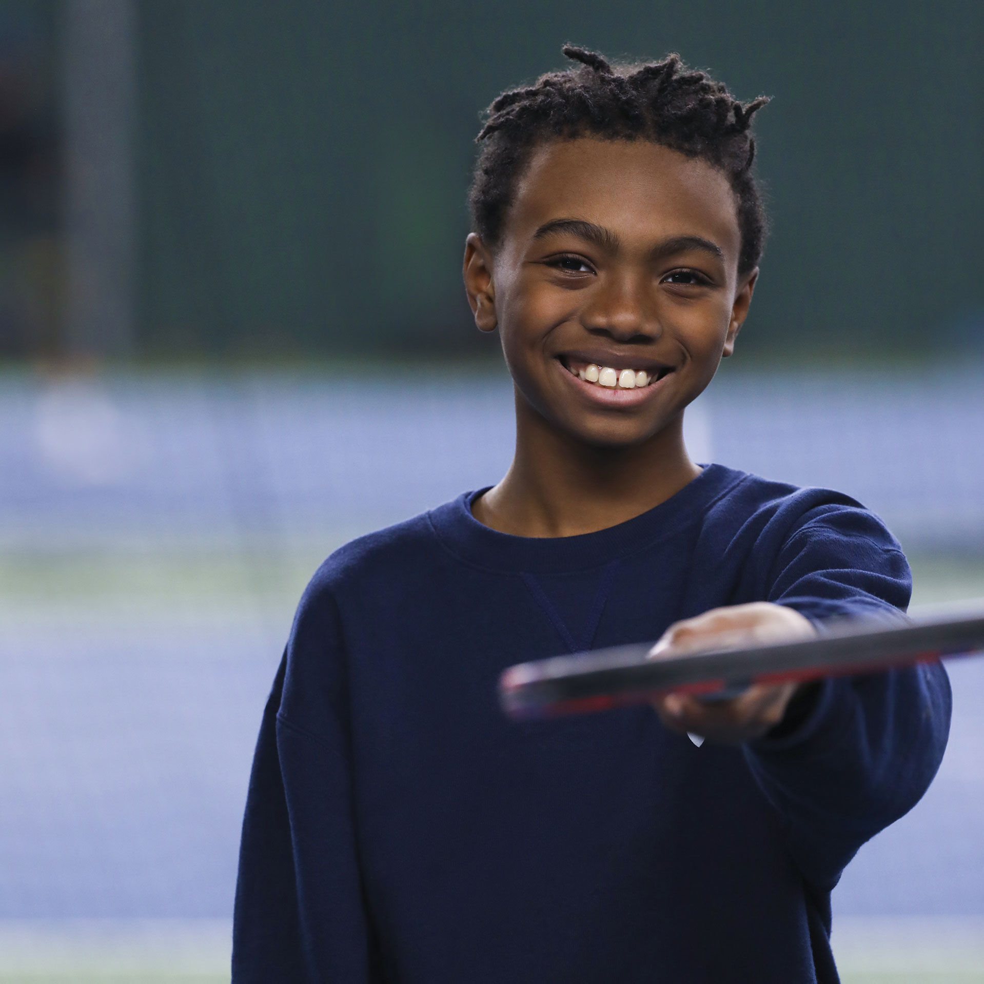 Un adolescent à la peau foncée et d'apparence masculine tenant une raquette de tennis souriant à la caméra.