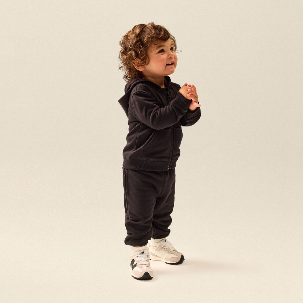 Tout-petit portant un ensemble molletonn&eacute; noir &agrave; capuchon et des chaussures de sport New Balance blanches, debout dans un d&eacute;cor de studio &eacute;pur&eacute;.