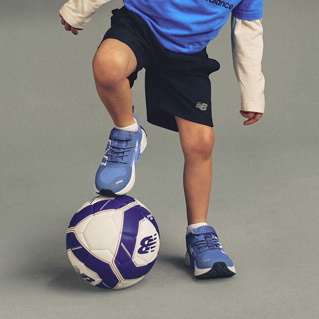 Un pied est pos&eacute; sur un ballon de soccer blanc et violet portant le logo New Balance bien visible, tandis que l'autre pied repose sur le sol. 