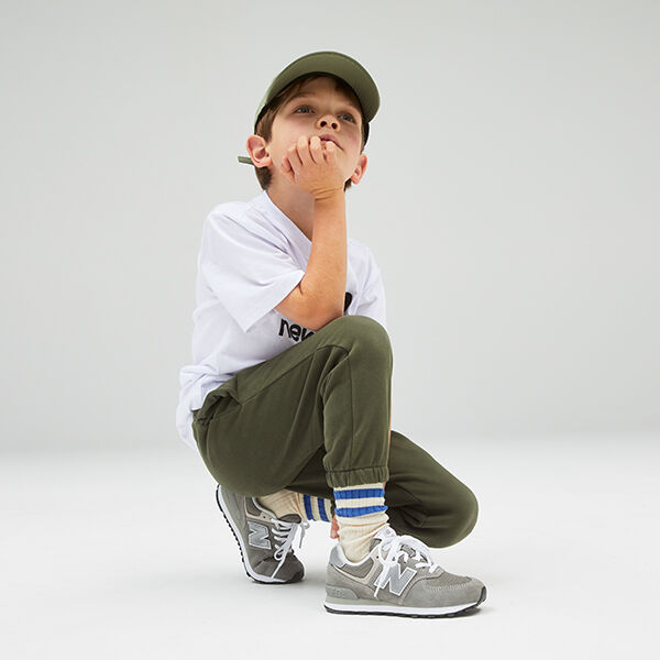 Un jeune garçon, accroupi devant un fond de studio gris clair, portant un tee-shirt NB Essentials blanc, un jogging vert olive et des chaussures de sport NB Classic 574 grises 
