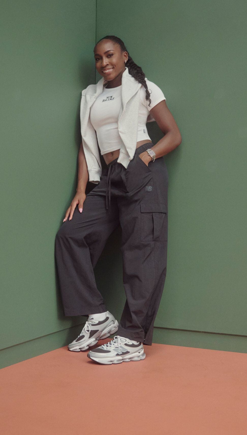 Coco Gauff appuy&eacute;e contre un mur vert d&rsquo;angle, v&ecirc;tue d&rsquo;un crop top, d&rsquo;un pantalon cargo large et de baskets New Balance blanches et grises.