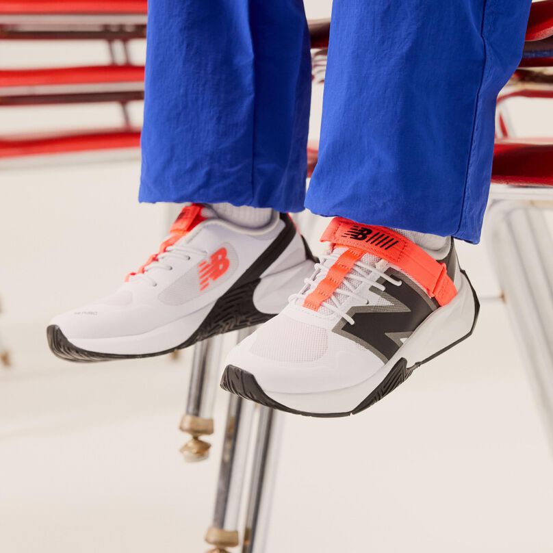 Jeune enfant tenant une pomme devant des chaises, avec un pantalon bleu et des chaussures blanches.