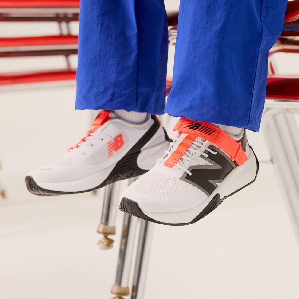 Jeune enfant tenant une pomme devant des chaises, avec un pantalon bleu et des chaussures blanches.