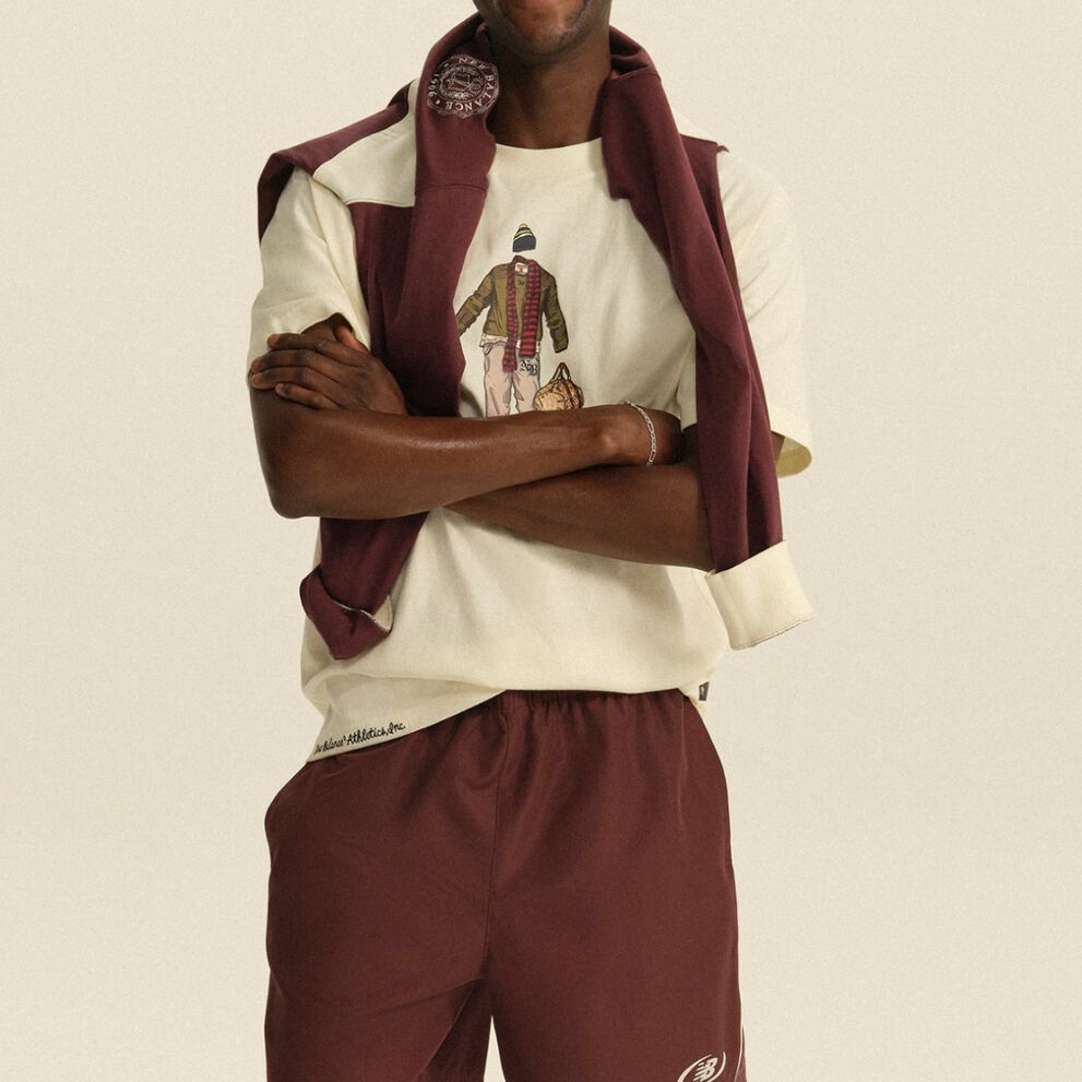Person wearing a cream graphic T-shirt with a burgundy sweatshirt draped over the shoulders and matching athletic pants in a clean studio setting.