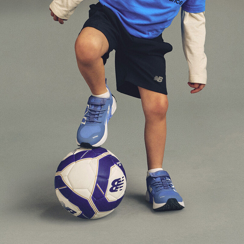 Un pied est pos&eacute; sur un ballon de soccer blanc et violet portant le logo New Balance bien visible, tandis que l'autre pied repose sur le sol. 