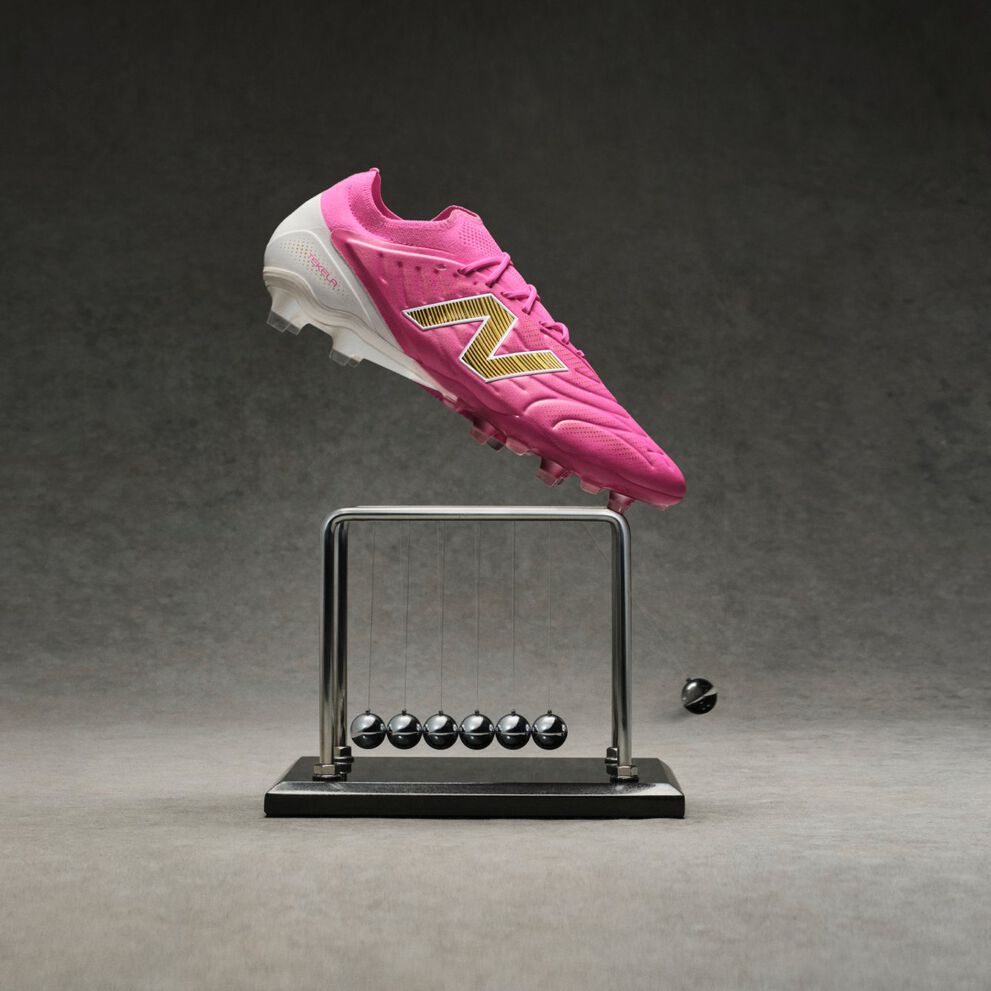 Bright pink New Balance soccer cleat with a metallic gold &ldquo;N&rdquo; logo appearing suspended above a black Newton&rsquo;s cradle on a gray studio surface, with one metal ball displaced to the side against a dark textured background.