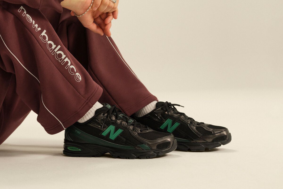 Close-up of black New Balance athletic sneakers with green accents paired with burgundy track pants and white crew socks in a clean studio setting.