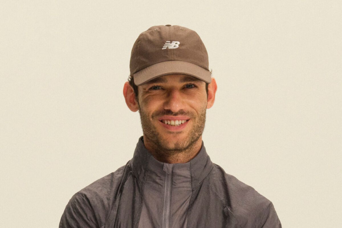 Person wearing a brown New Balance cap and a gray color-block performance jacket in a clean studio setting.