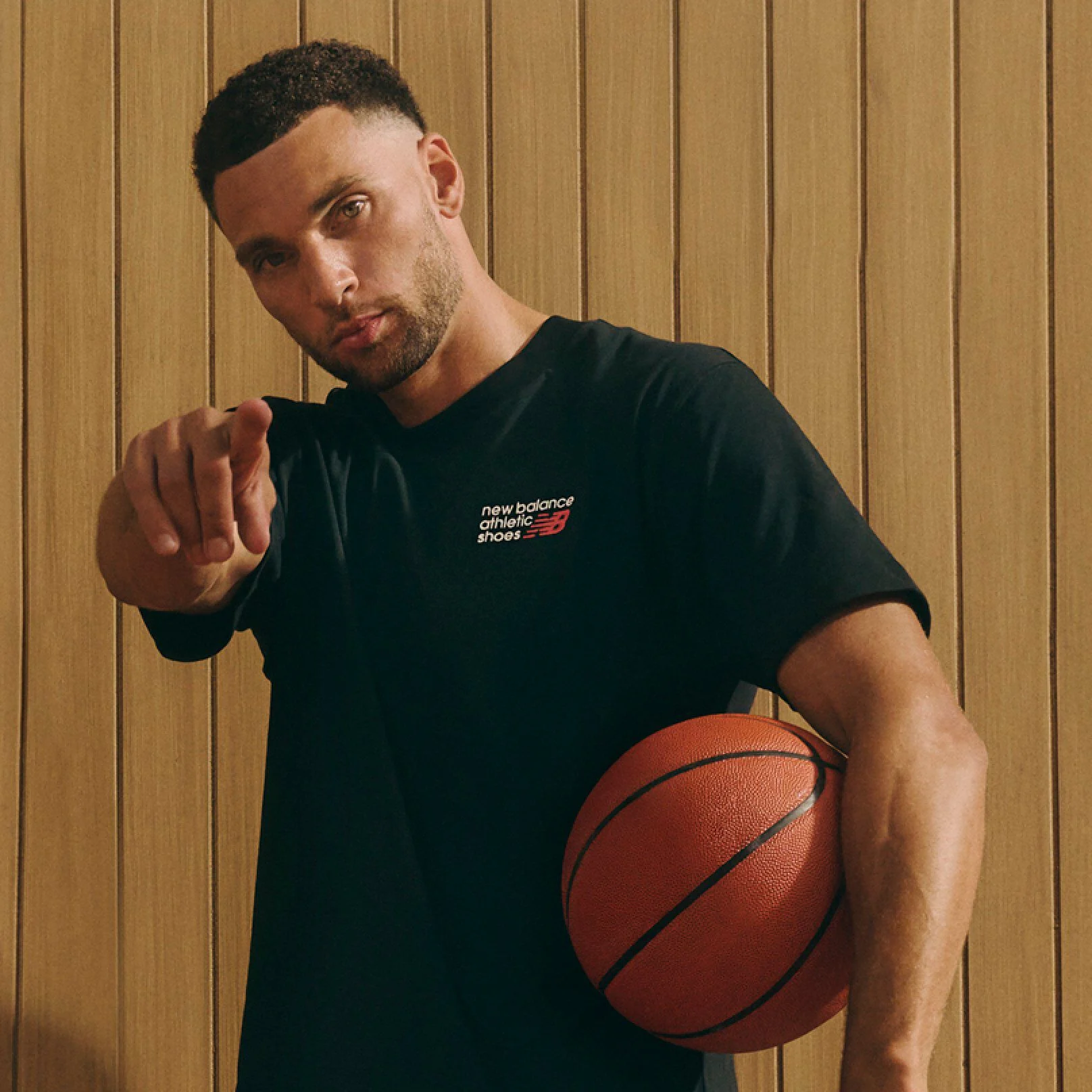 Une personne portant un t-shirt de sport noir New Balance pointe vers la cam&eacute;ra tout en tenant un ballon de basket sur le c&ocirc;t&eacute;, debout devant un fond en panneaux de bois.