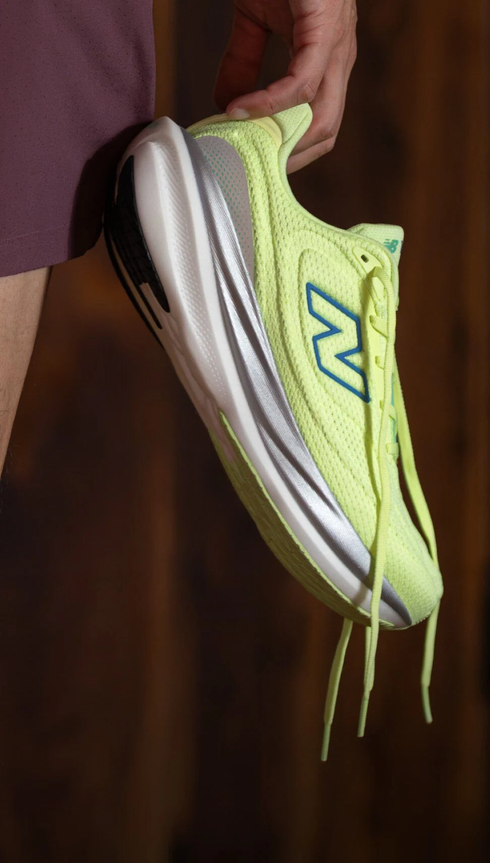 Gros plan d&rsquo;une chaussure de course New Balance jaune-vert vif avec logo bleu et semelle blanche, tenue par une personne portant un short bordeaux.&nbsp;