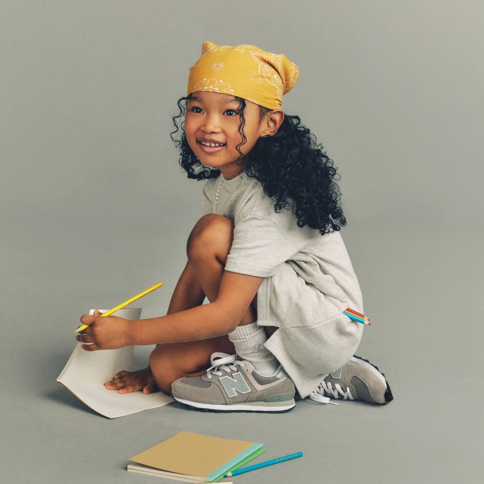 Child crouching on the floor writing in a notebook with colored pencils, wearing a yellow bandana, gray outfit, white socks, and gray New Balance sneakers against a plain background.