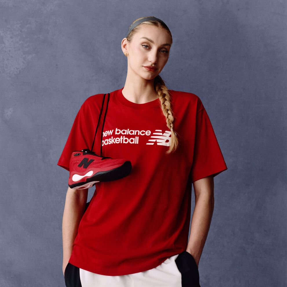 Athlete wearing a red New Balance basketball T-shirt holding a matching red and black sneaker against a gray studio background, with the &lsquo;new balance basketball&rsquo; logo visible on the shirt.