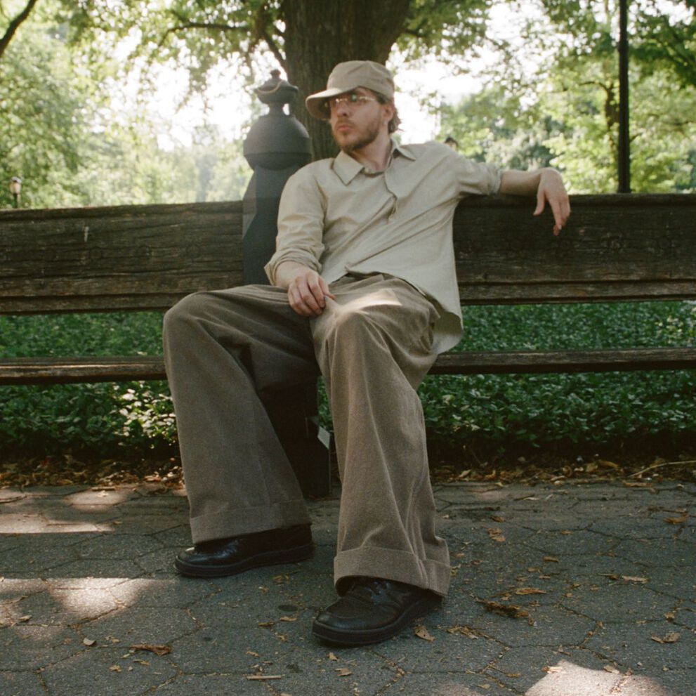 Jack Harlow reclining on a bench in a park wearing dark brown New Balance sneakers.